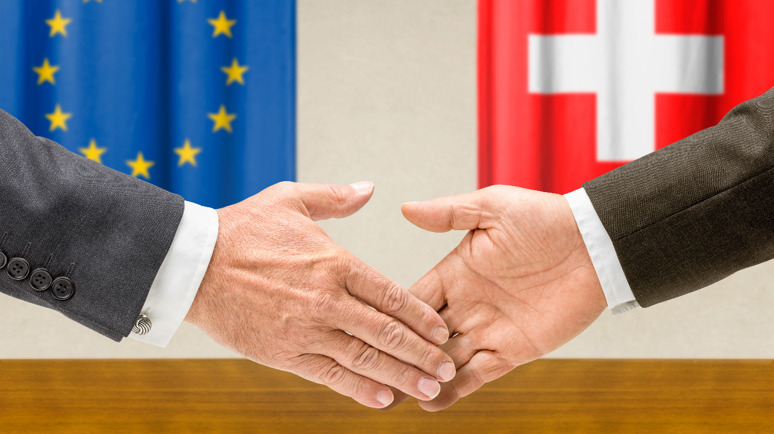 Zwei Hände geben sich die Hand. Im Hintergrund sind eine EU Flagge und eine Schweizer Flagge